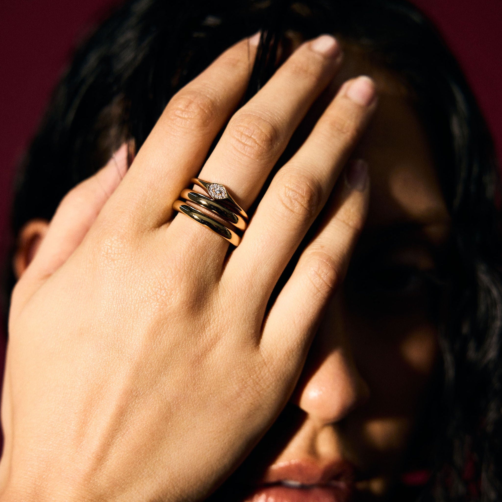 Person covering face with hand wearing gold rings and bracelet against a dark background