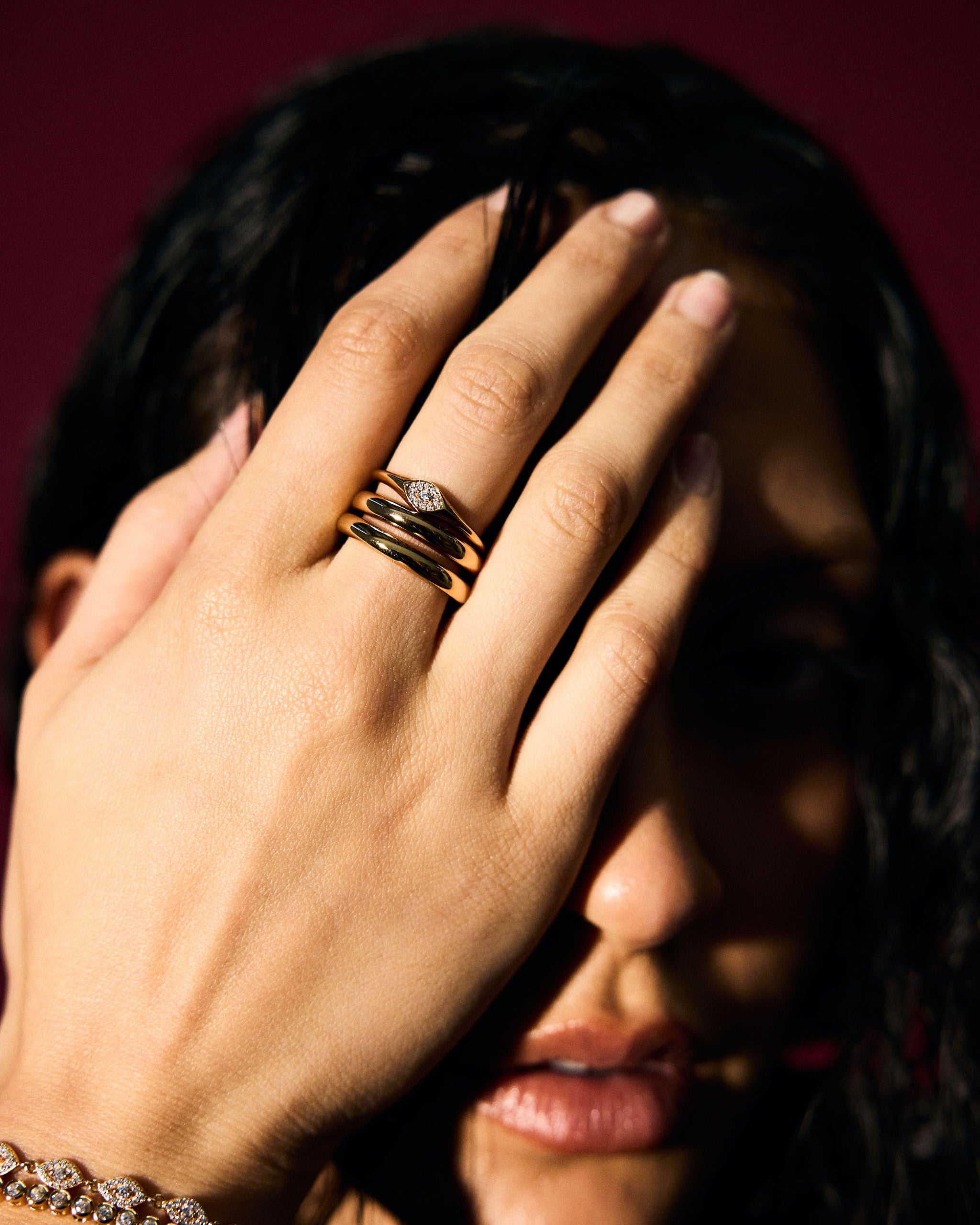 Person covering face with hand wearing gold rings and bracelet against a dark background