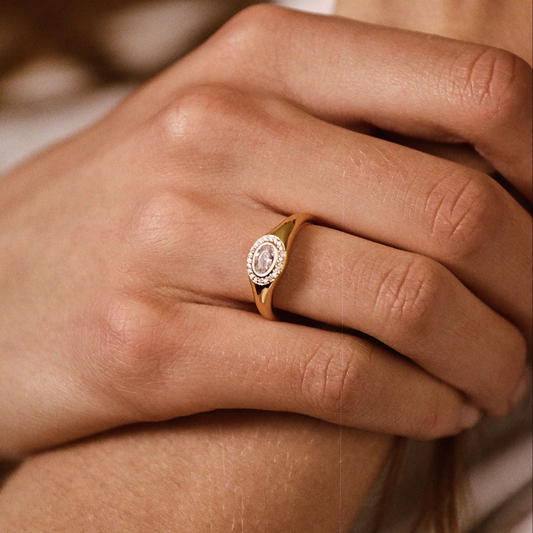 A close-up lifestyle shot of a person's hand resting on their arm, wearing a delicate gold ring with a round bezel-set white gemstone.