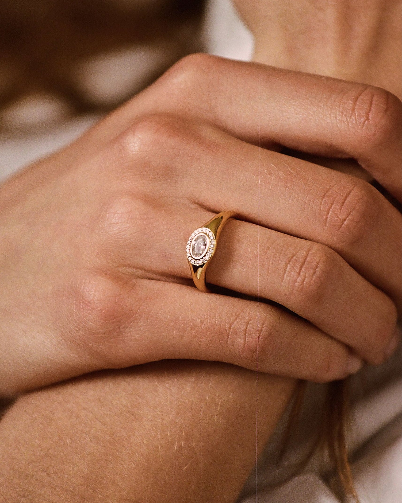 A close-up lifestyle shot of a person's hand resting on their arm, wearing a delicate gold ring with a round bezel-set white gemstone.
