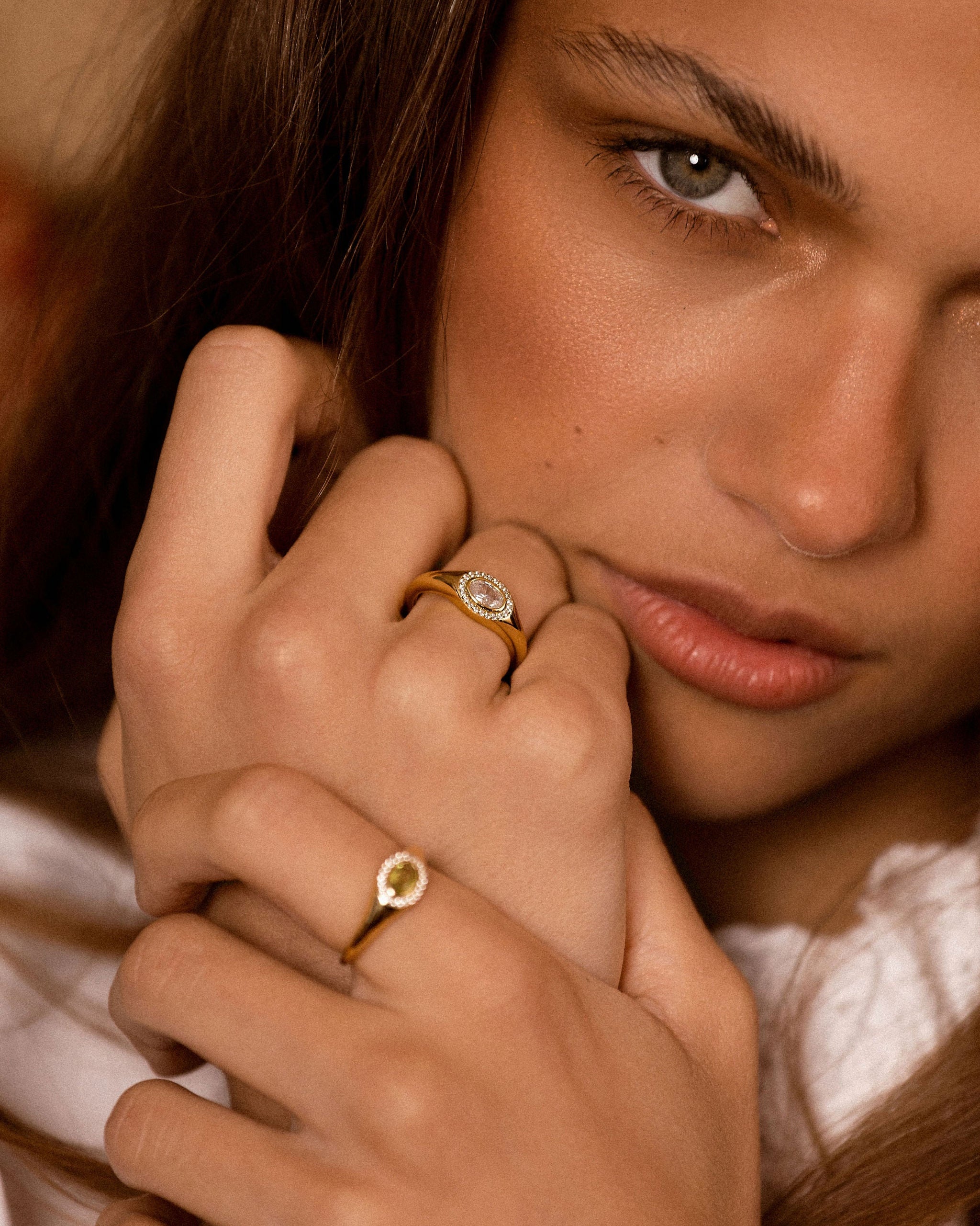 A close-up lifestyle shot of a woman wearing multiple gold rings with small clear gemstones on her fingers, set against a warm, softly blurred background.