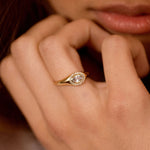 A close-up lifestyle shot of a woman wearing a gold ring with a clear round-cut center stone and a halo of small clear stones.