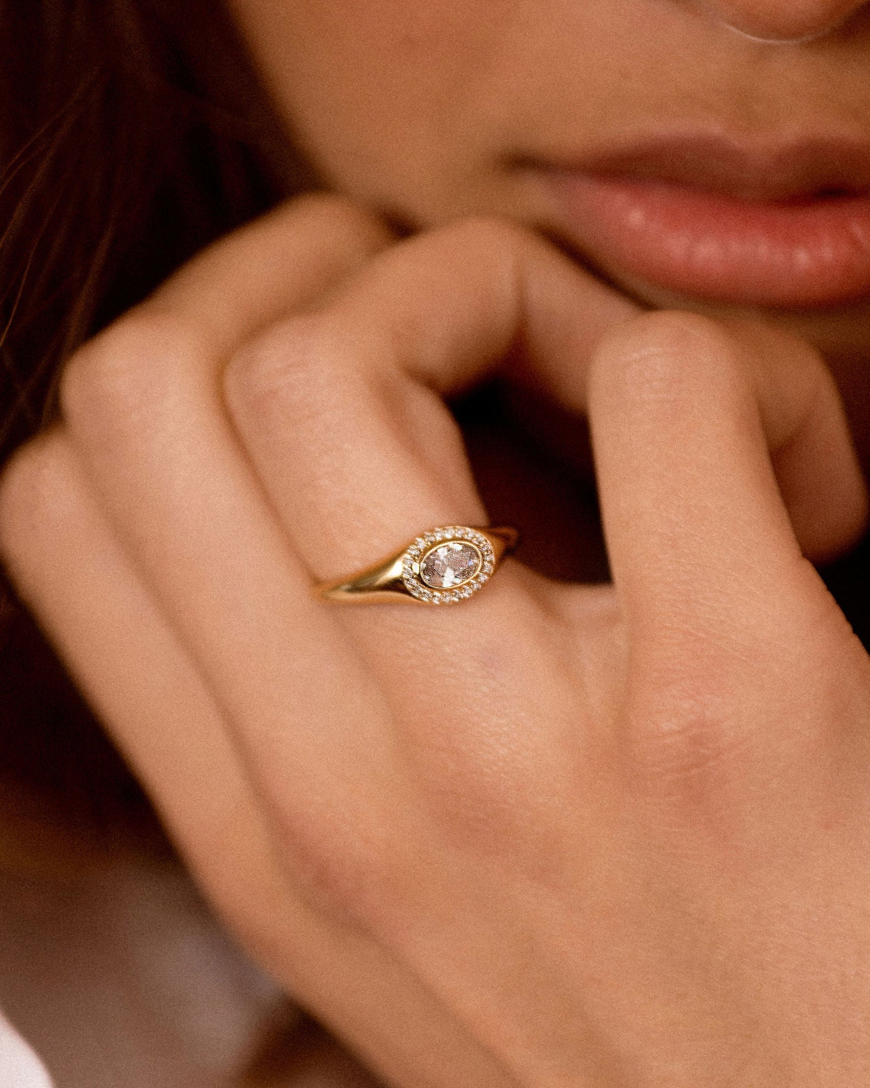 A close-up lifestyle shot of a woman wearing a gold ring with a clear round-cut center stone and a halo of small clear stones.