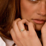A close-up lifestyle shot of a woman wearing a delicate gold ring with a central oval-cut clear gemstone on her finger.