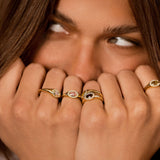 A close-up lifestyle shot of a model wearing five gold rings on their fingers, with their hands covering their mouth. The rings feature various designs including a heart, an eye, and crescent moon motifs, set with clear stones.