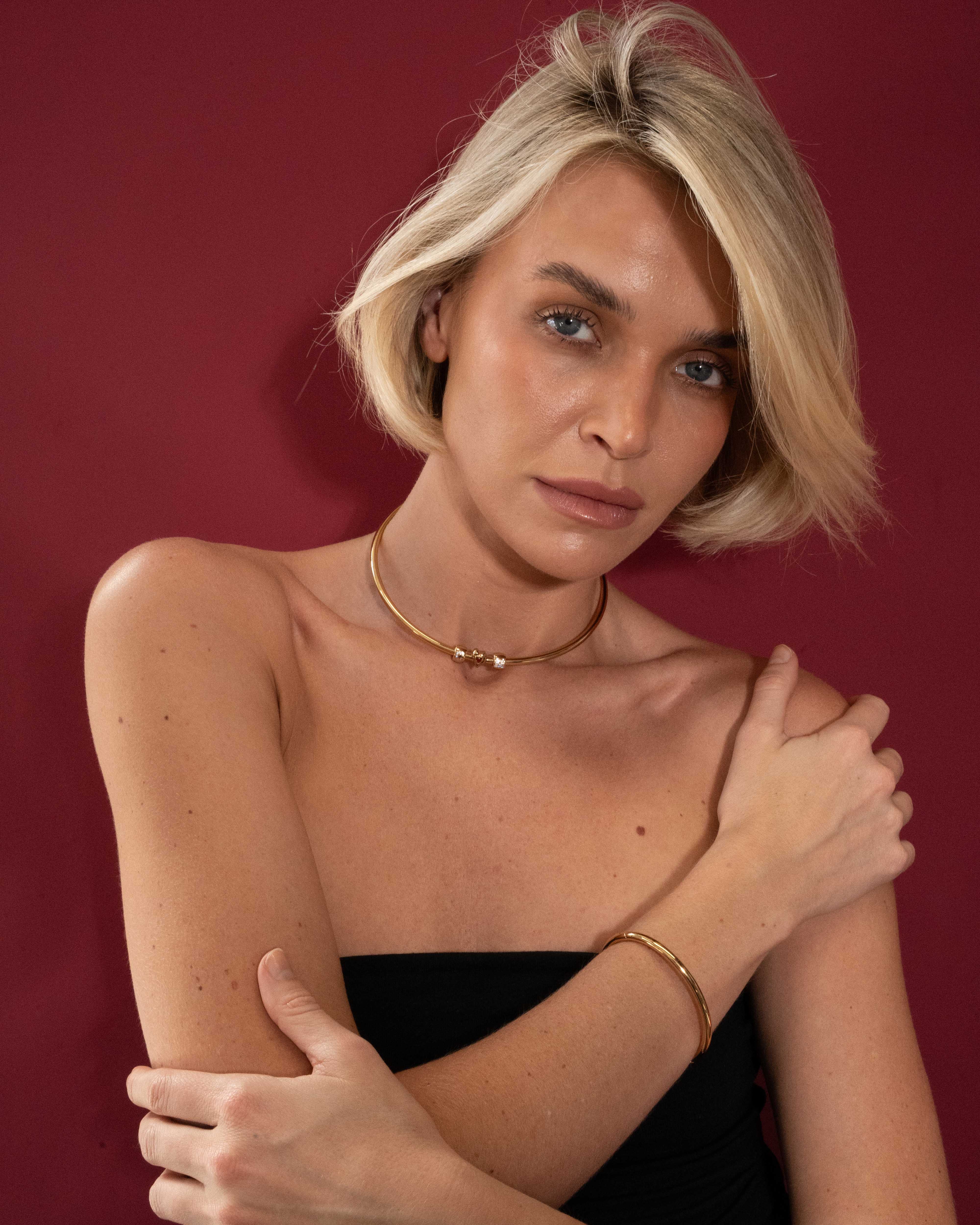 Woman with short blonde hair wearing a black top and gold jewelry against a red background