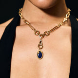 Close-up of a person wearing a gold necklace with a blue pendant against a black background