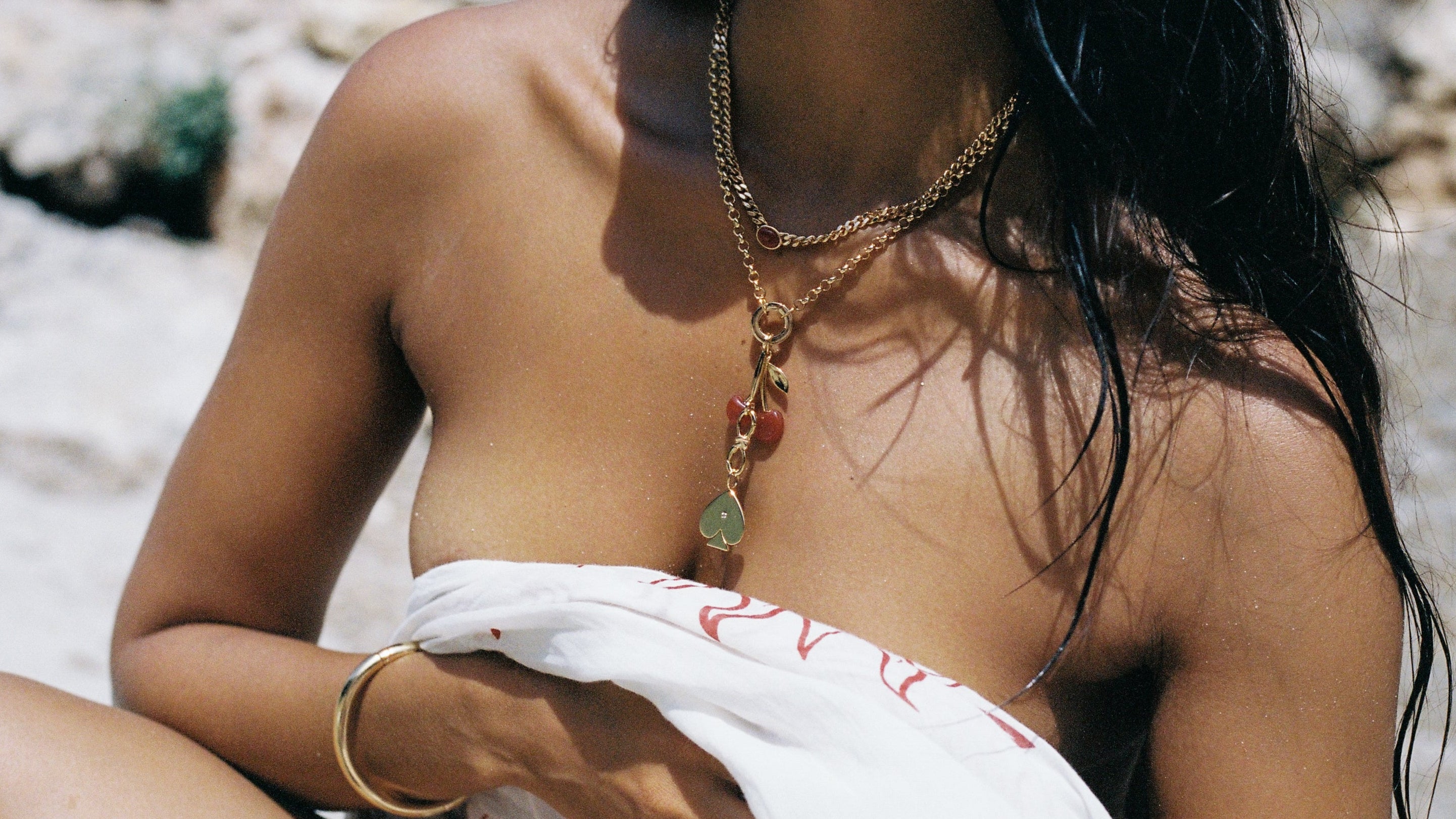 Person sitting on a rocky surface with a white garment draped over their shoulders, wearing gold jewelry.