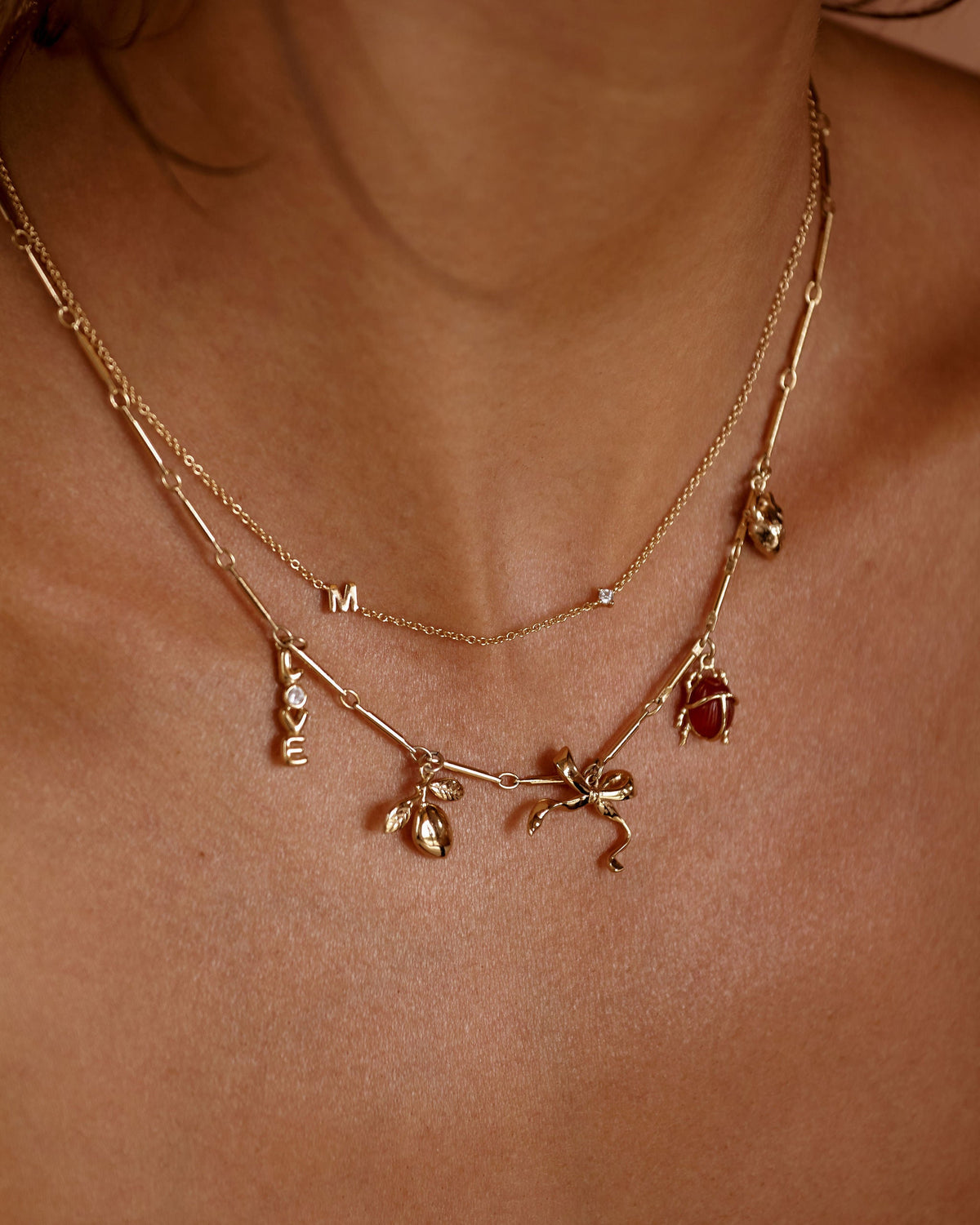 A close-up of a person's neck and chest wearing a layered gold necklace with small dangling charms, including a dragonfly and a red gemstone accent.