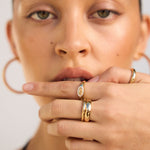 Close-up of a woman's face with gold rings on her fingers against a neutral background