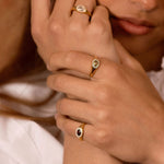 A close-up lifestyle shot of a model wearing three gold rings with oval-cut stones in different colors (clear, light green, and dark red) on her fingers.