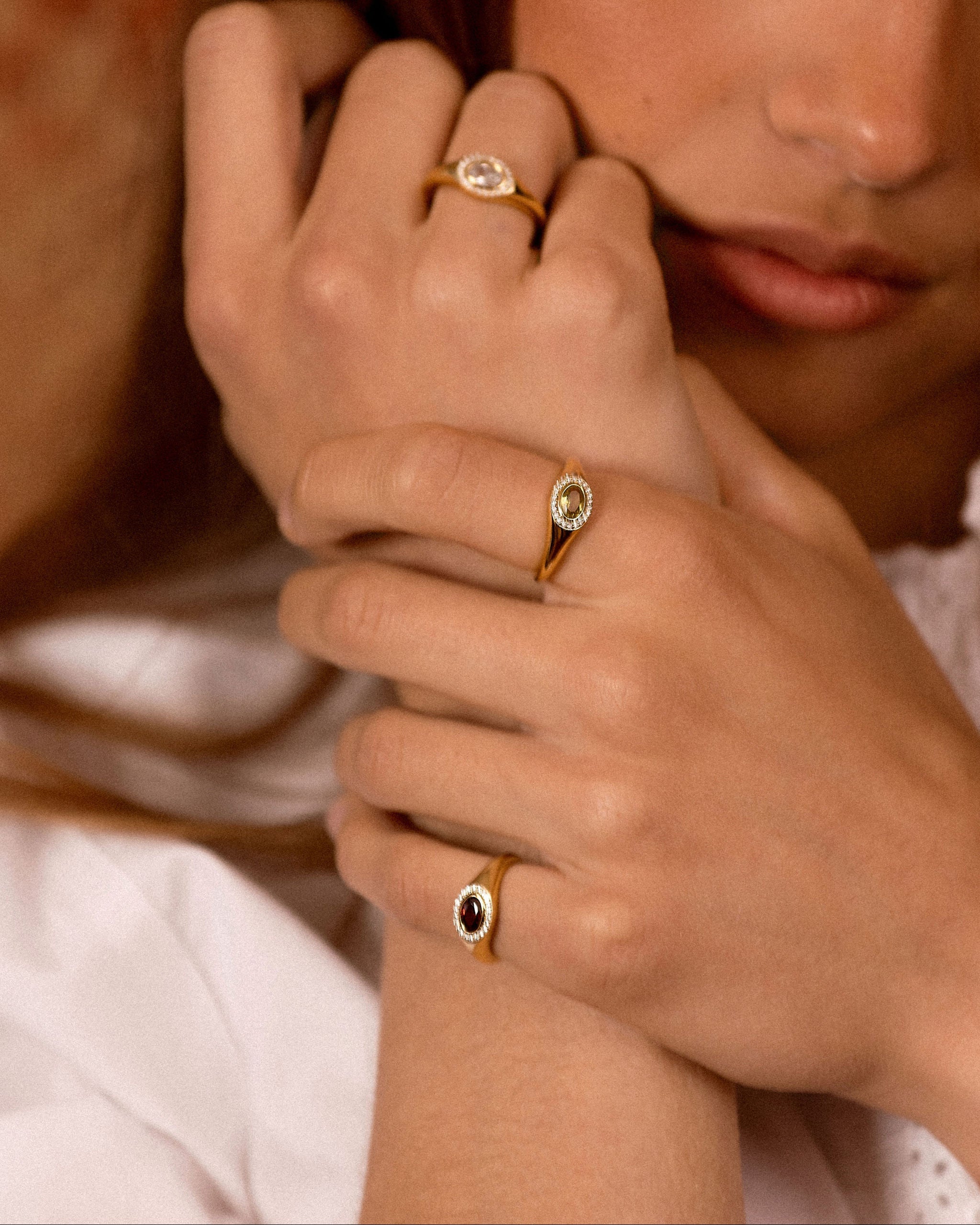 A close-up lifestyle shot of a model wearing three gold rings with oval-cut stones in different colors (clear, light green, and dark red) on her fingers.
