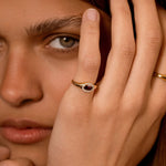 A close-up lifestyle shot of a model wearing a gold ring with a dark red oval stone and small clear stones, with her hand resting near her face.