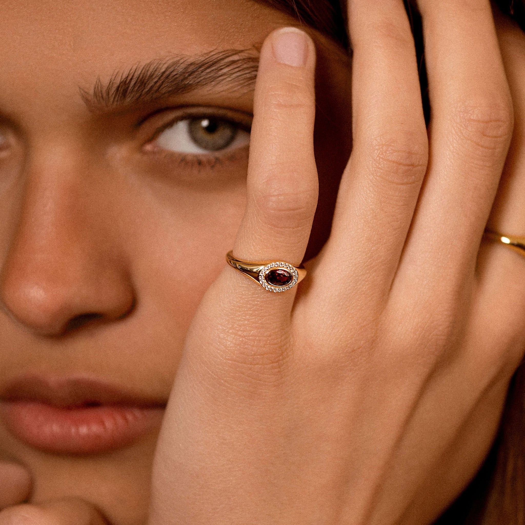 A close-up lifestyle shot of a model wearing a gold ring with a dark red oval stone and small clear stones, with her hand resting near her face.