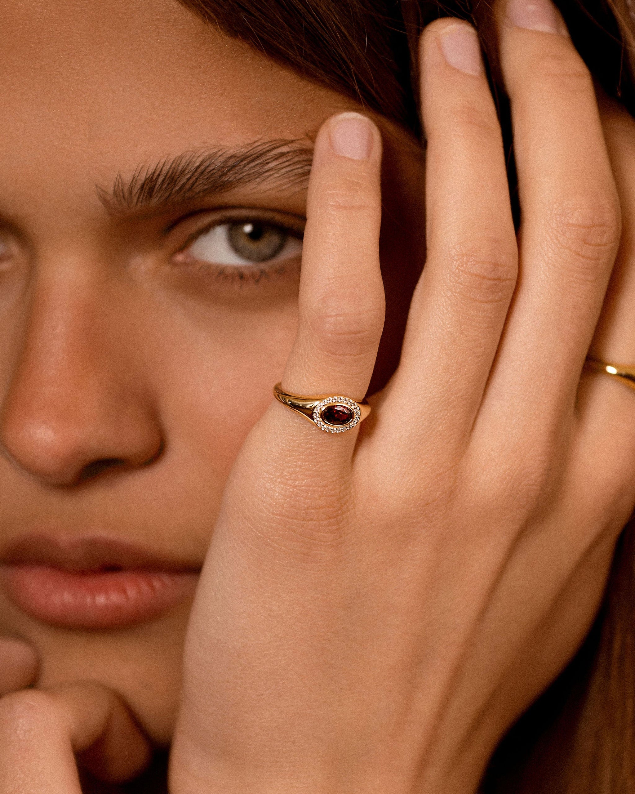 A close-up lifestyle shot of a model wearing a gold ring with a dark red oval stone and small clear stones, with her hand resting near her face.