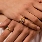 A close-up of a person's hands wearing two gold rings. The larger ring on the middle finger features a double-row setting with red gemstones, and the smaller ring on the ring finger has a single green gemstone.