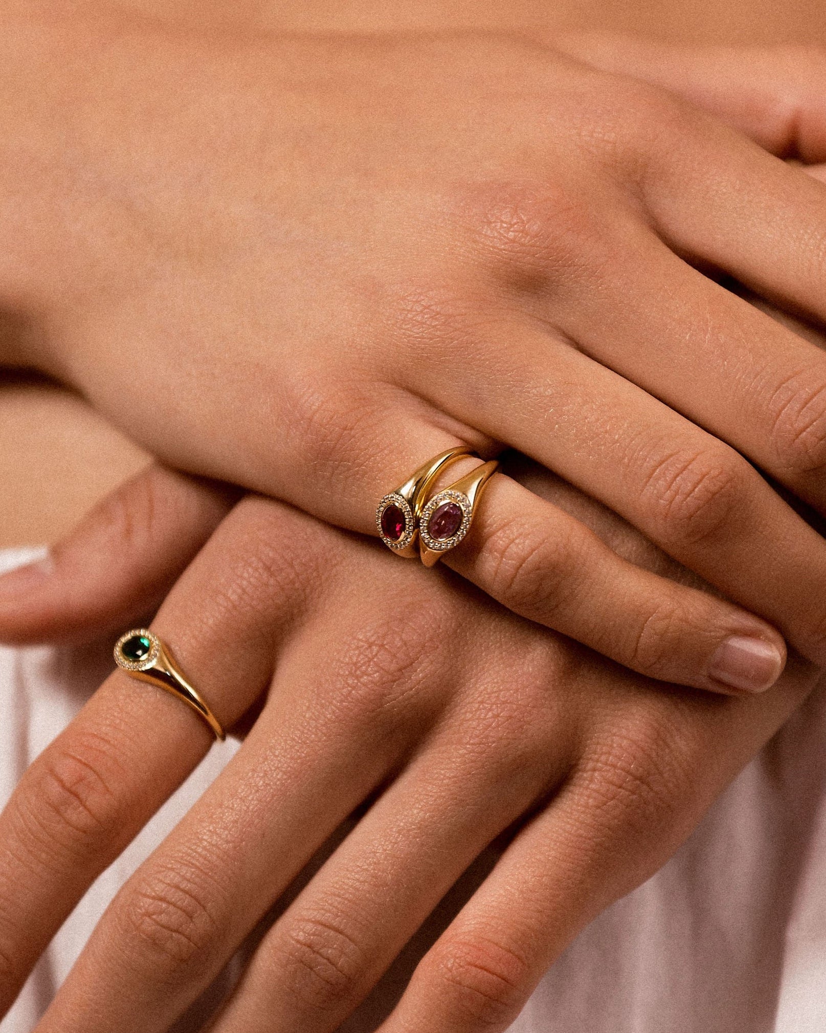 A close-up of a person's hands wearing two gold rings. The larger ring on the middle finger features a double-row setting with red gemstones, and the smaller ring on the ring finger has a single green gemstone.