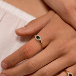 A close-up of a person's hands resting on white fabric, showcasing two gold rings with oval gemstones. One ring features a dark green stone, and the other features a red stone.