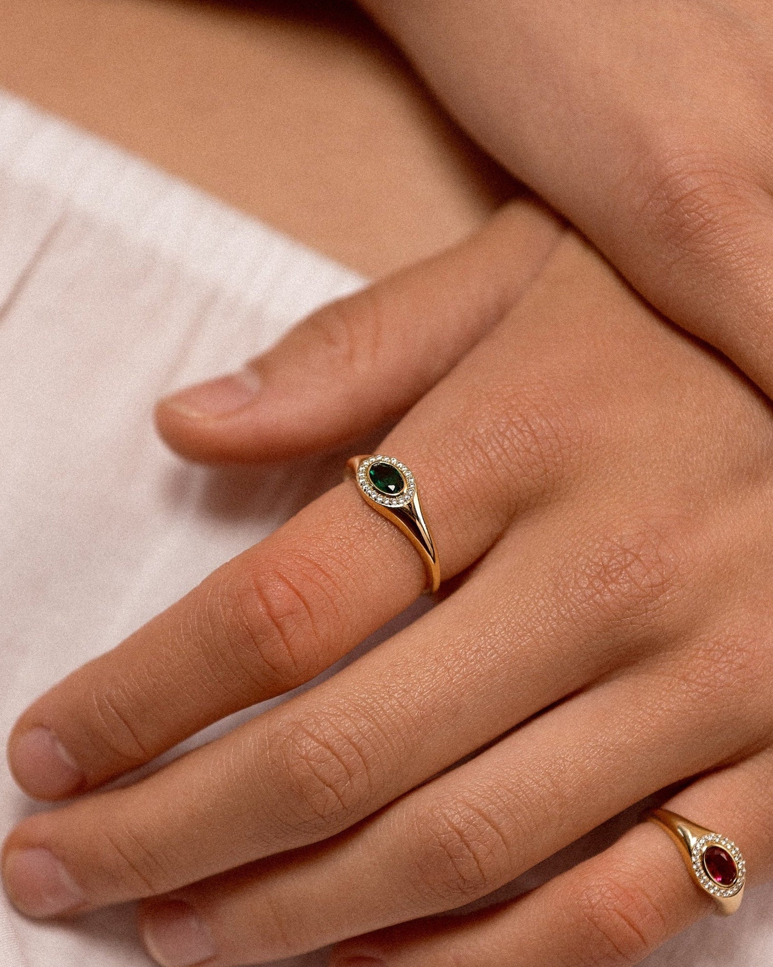 A close-up of a person's hands resting on white fabric, showcasing two gold rings with oval gemstones. One ring features a dark green stone, and the other features a red stone.