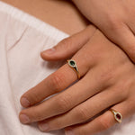 A close-up of a person's hands resting on white fabric, showcasing two gold rings with oval gemstones. One ring features a dark green stone, and the other features a red stone.