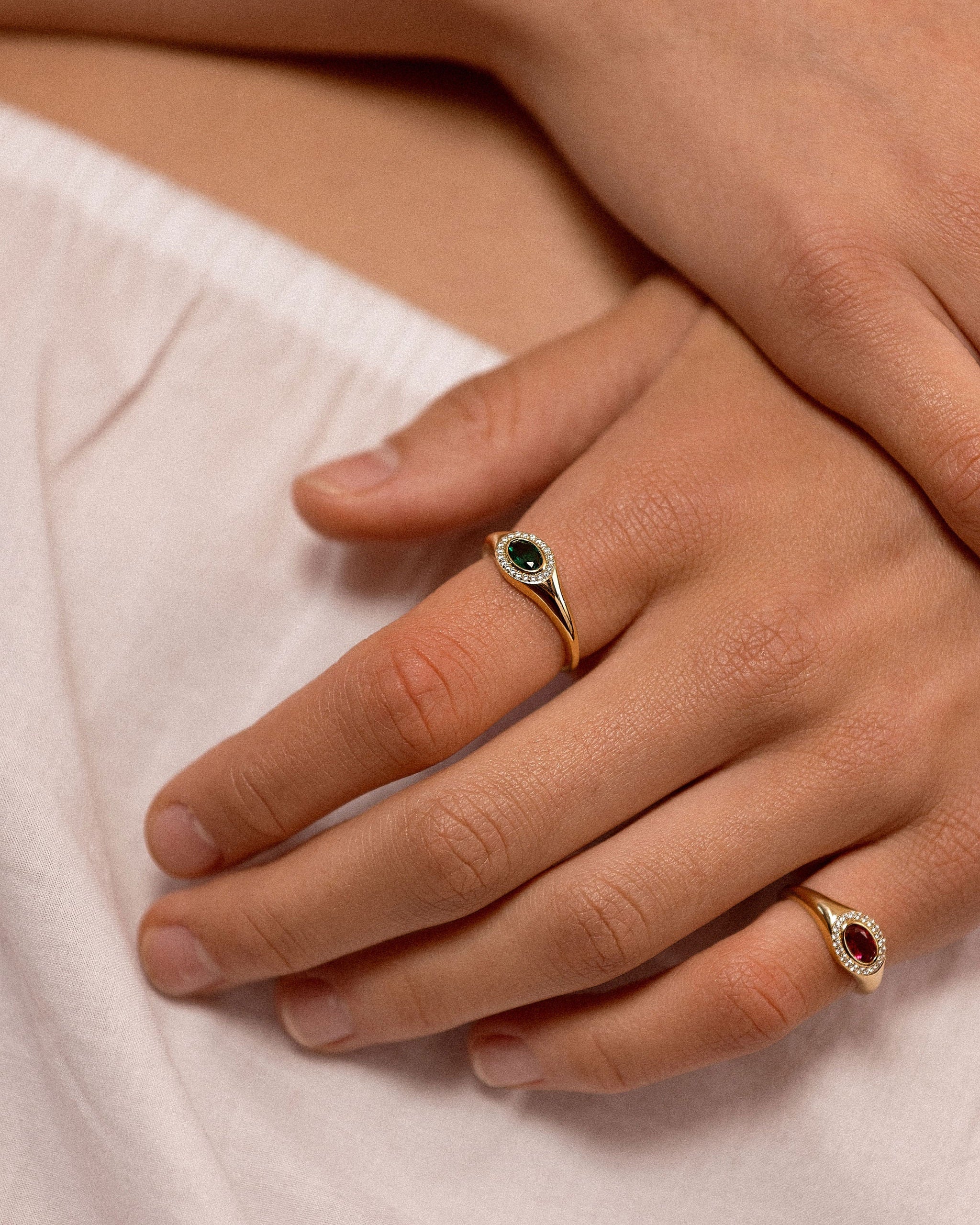 A close-up of a person's hands resting on white fabric, showcasing two gold rings with oval gemstones. One ring features a dark green stone, and the other features a red stone.