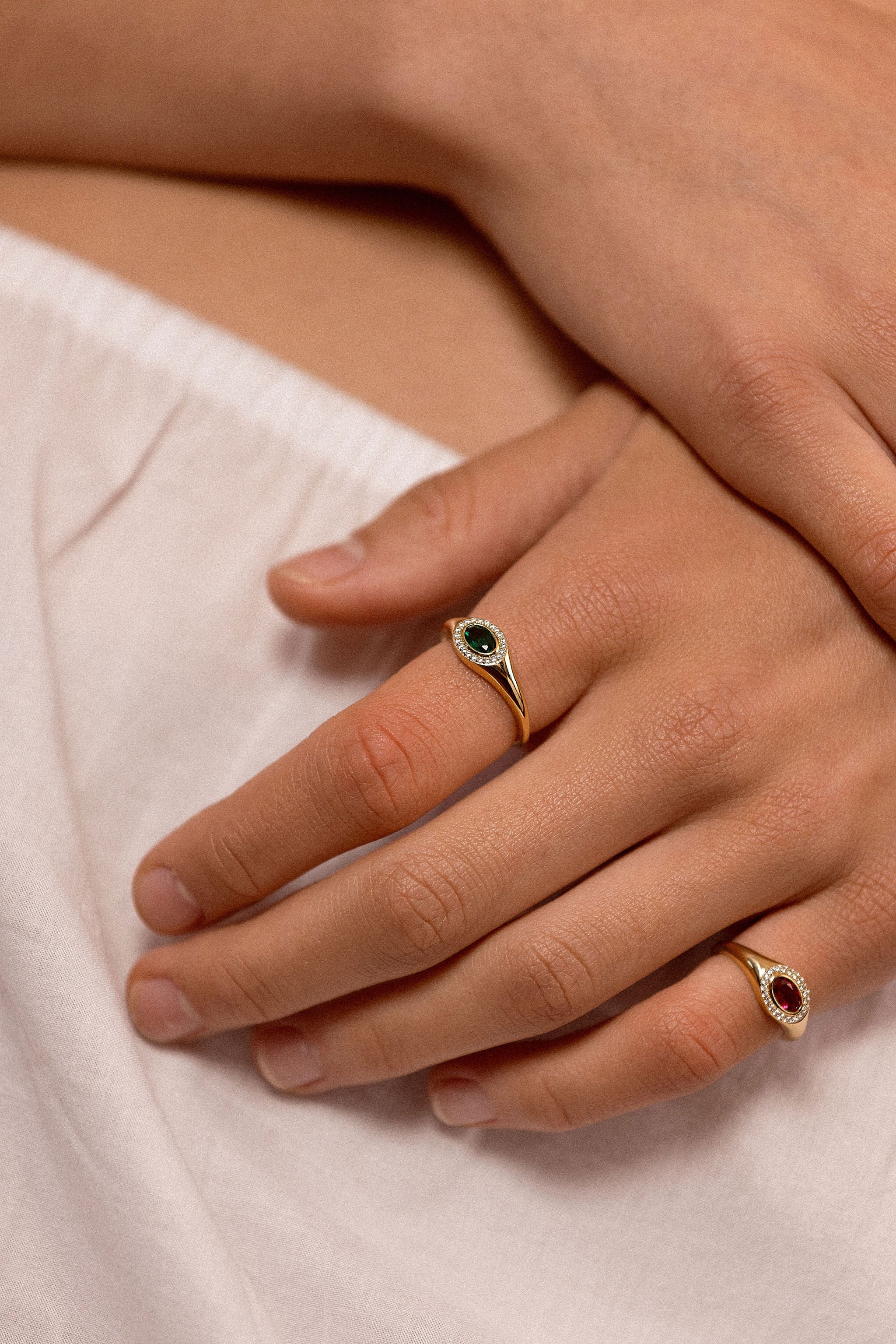 A close-up of a person's hands resting on white fabric, showcasing two gold rings with oval gemstones. One ring features a dark green stone, and the other features a red stone.