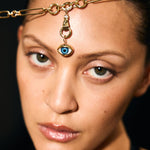 Woman wearing a gold headpiece with an eye pendant against a dark background