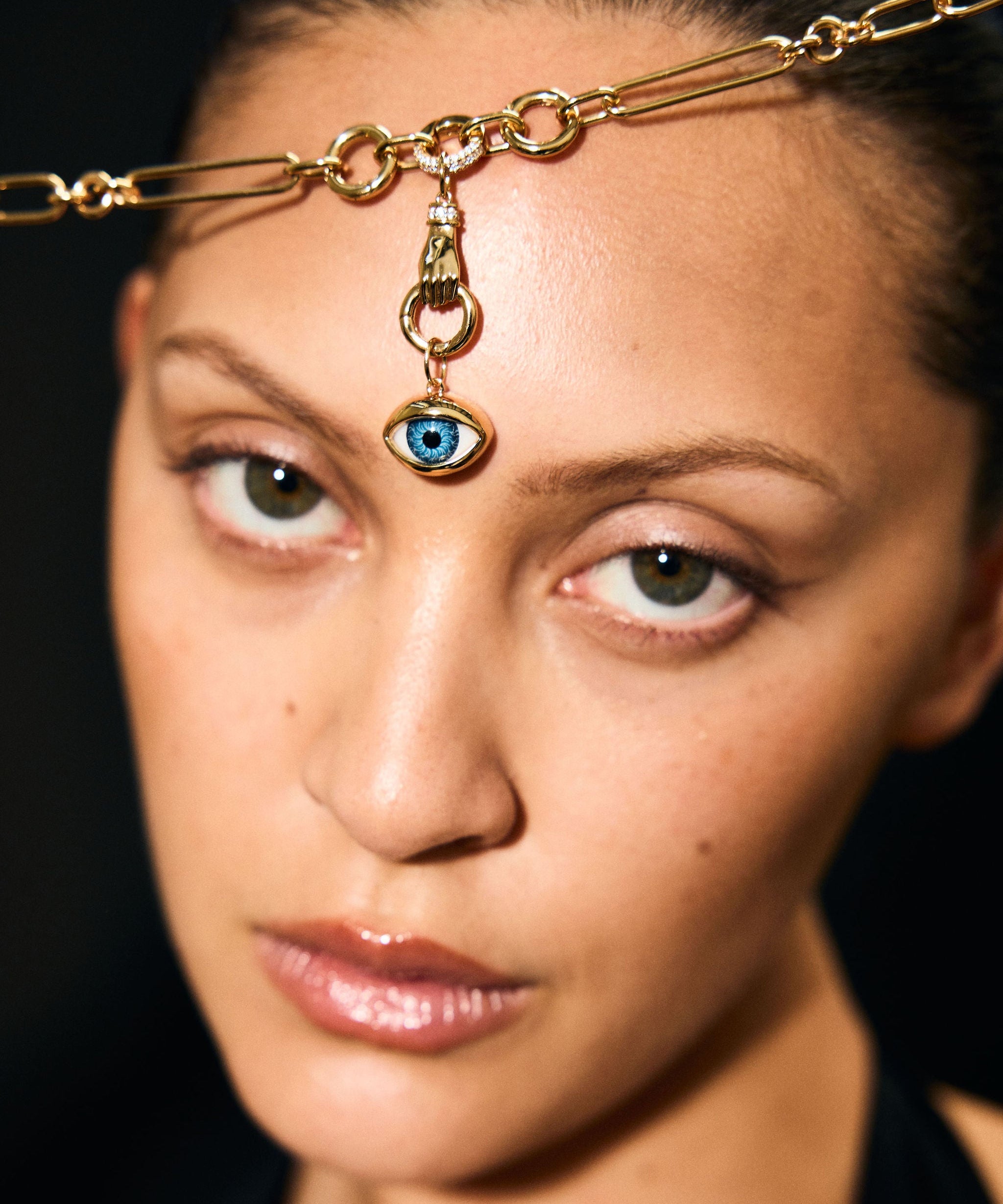 Woman wearing a gold headpiece with an eye pendant against a dark background