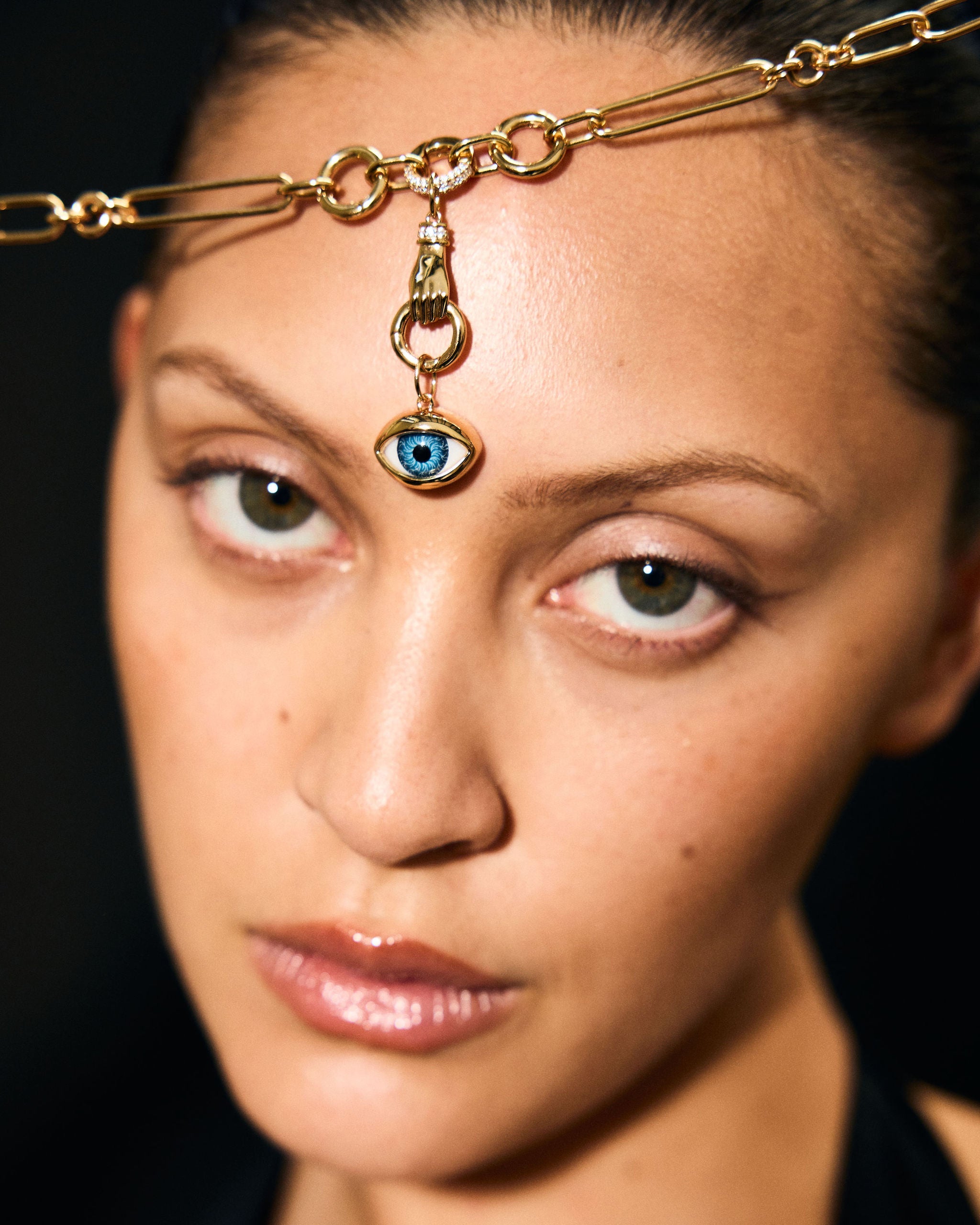 Woman wearing a gold headpiece with an eye pendant against a dark background