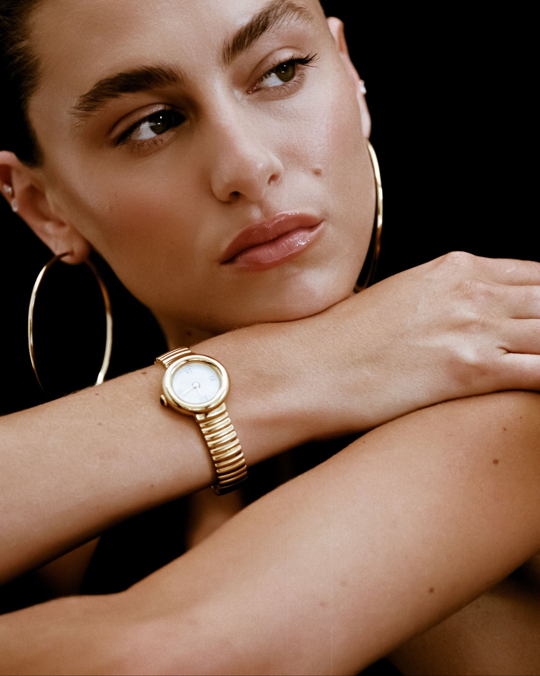 Woman wearing a gold watch and hoop earrings against a black background