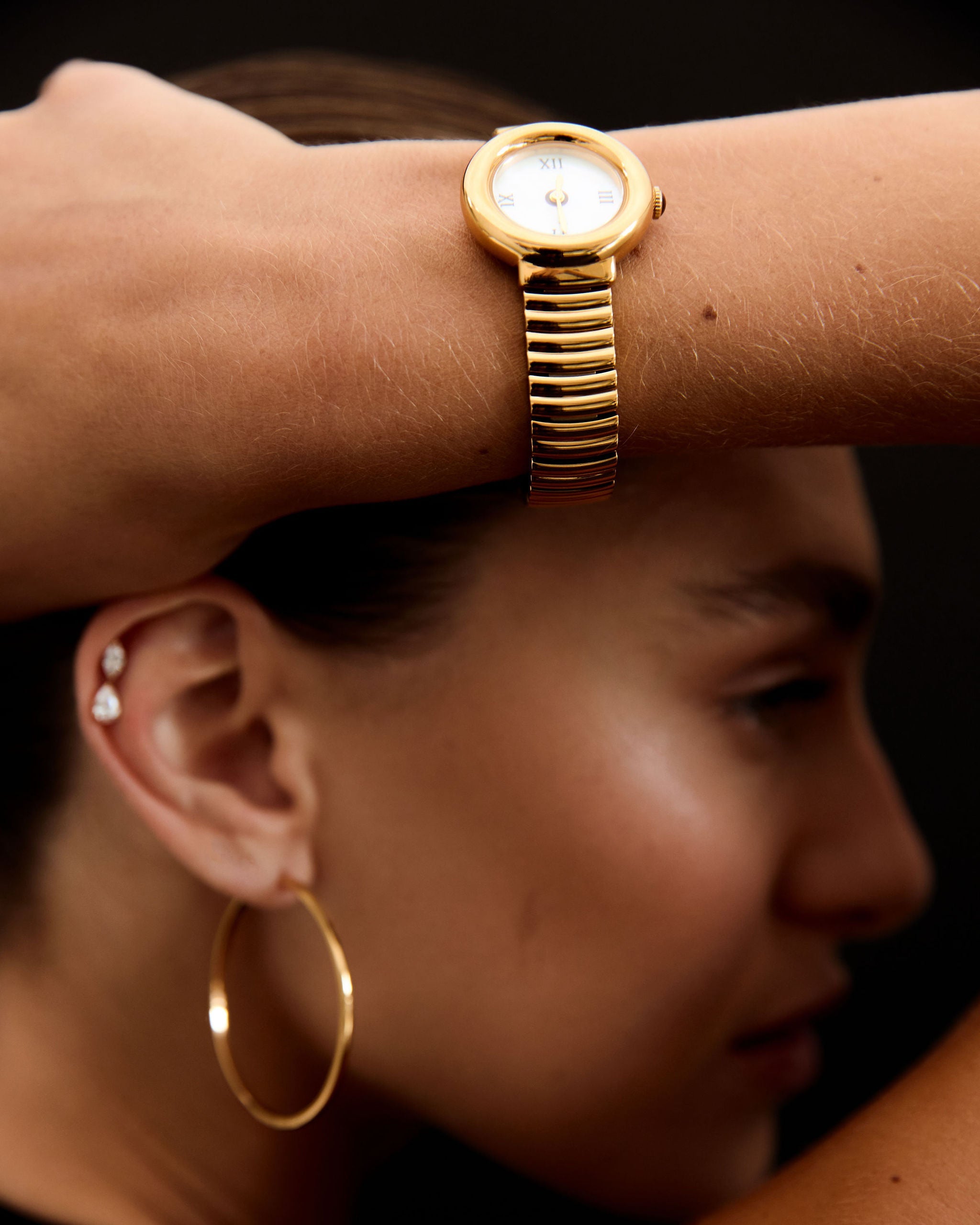 Close-up of a person wearing a gold watch and hoop earrings with a dark background