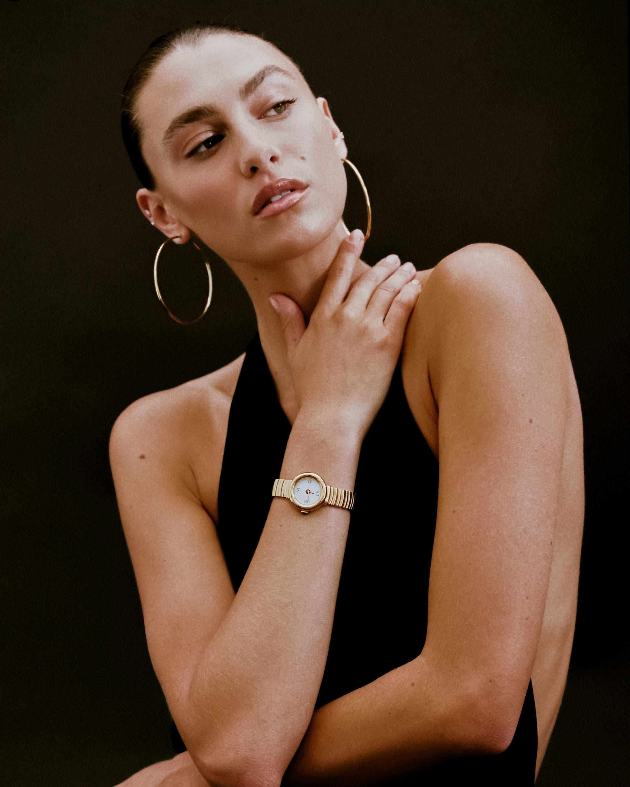 Woman wearing gold hoop earrings and a watch on a dark background