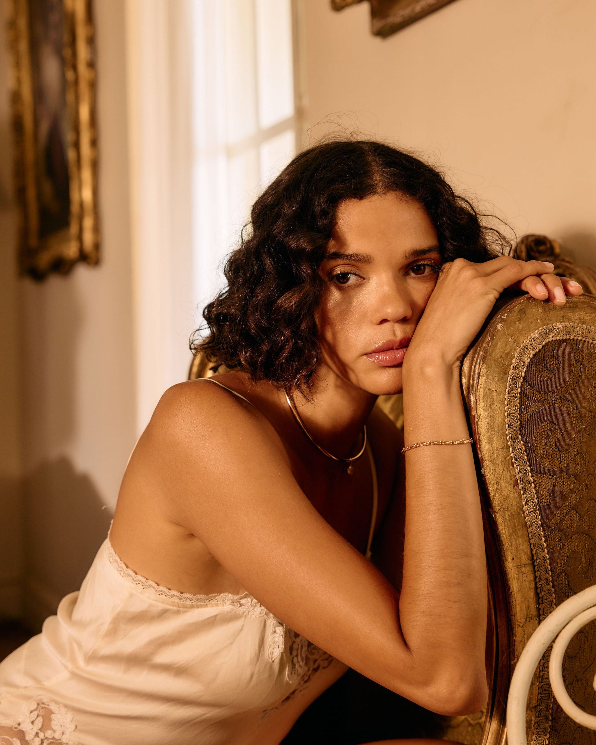 Woman in a white lace nightgown sitting on a chair in a warmly lit room.