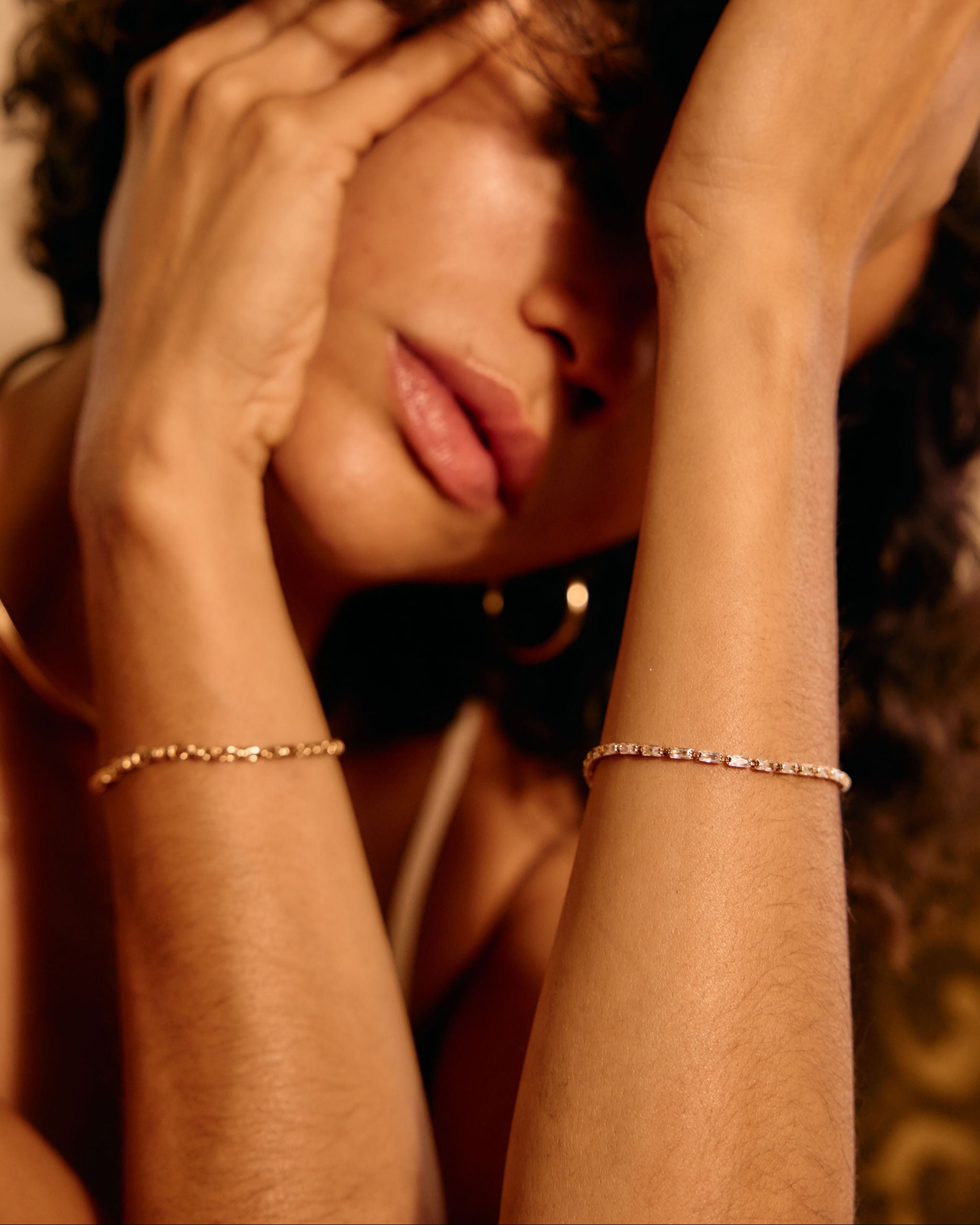 Woman with hands on head wearing gold bracelets in warm-toned lighting