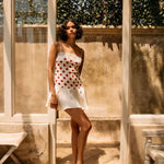 Woman in a polka dot dress standing in a sunlit outdoor setting with wooden beams and plants.