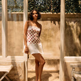 Woman in a polka dot dress standing in a sunlit outdoor setting with wooden beams and plants.