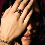 Person covering face with hand wearing gold rings and bracelet against a dark background