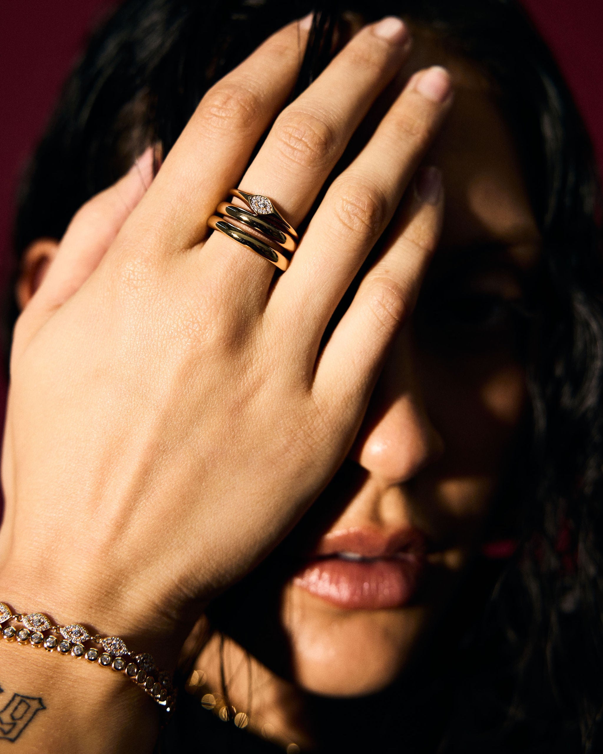 Person covering face with hand wearing gold rings and bracelet against a dark background