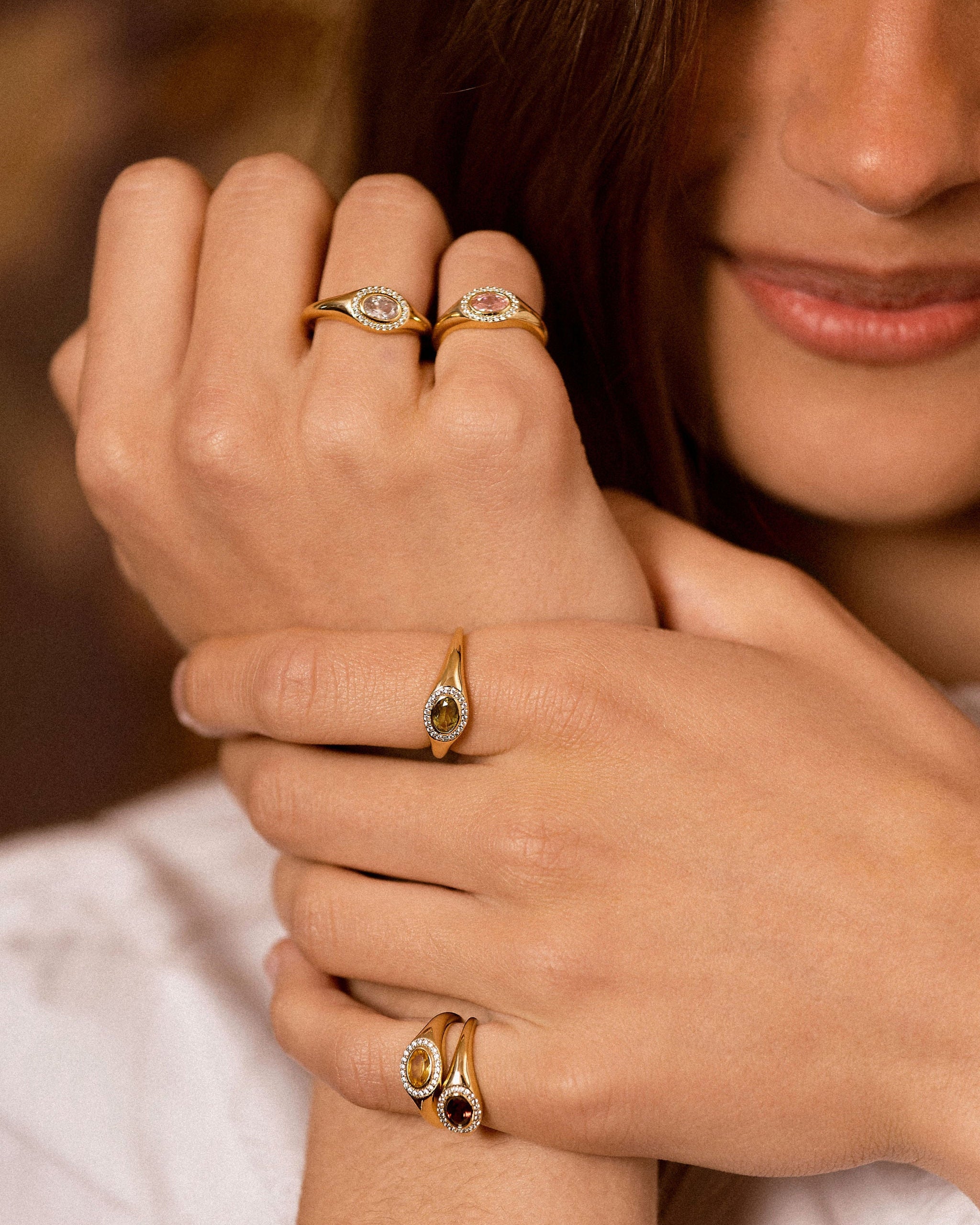 A close-up lifestyle shot of a model's hands wearing multiple gold rings with oval-cut stones, including a green stone and a dark red stone.