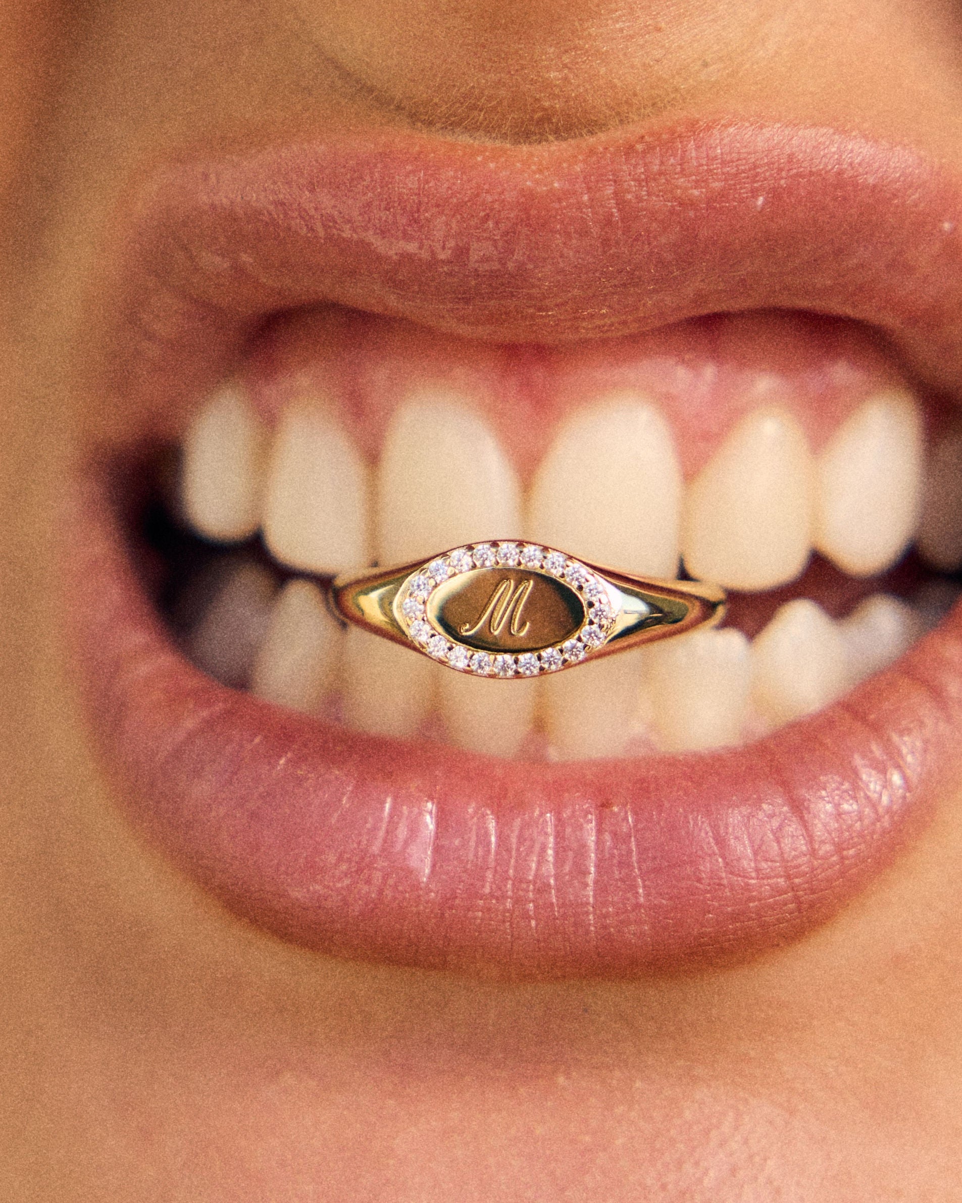 Close-up of a mouth wearing a gold ring with a letter 'M' on a blurred background
