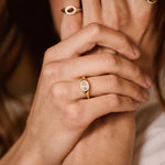 A close-up lifestyle shot of a person wearing several gold rings, with their hands covering part of their face. The rings feature oval and round settings with small colored stones, including a prominent gold ring with a round white stone on the ring finger.
