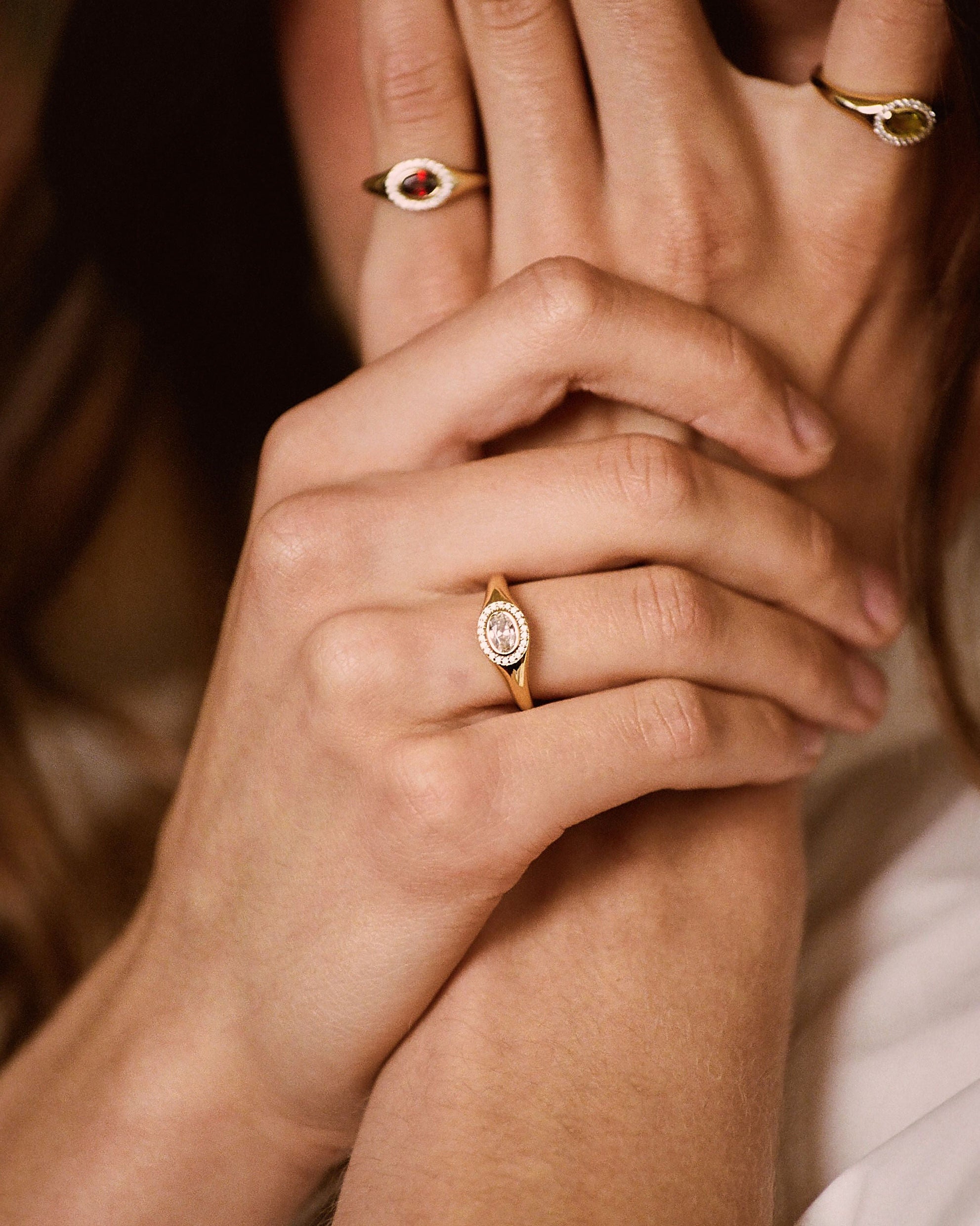 A close-up lifestyle shot of a person wearing several gold rings, with their hands covering part of their face. The rings feature oval and round settings with small colored stones, including a prominent gold ring with a round white stone on the ring finger.