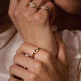 Close-up of a person wearing multiple gold rings with oval gemstones, including one with a pink stone and another with a dark stone, against a soft white lace garment.