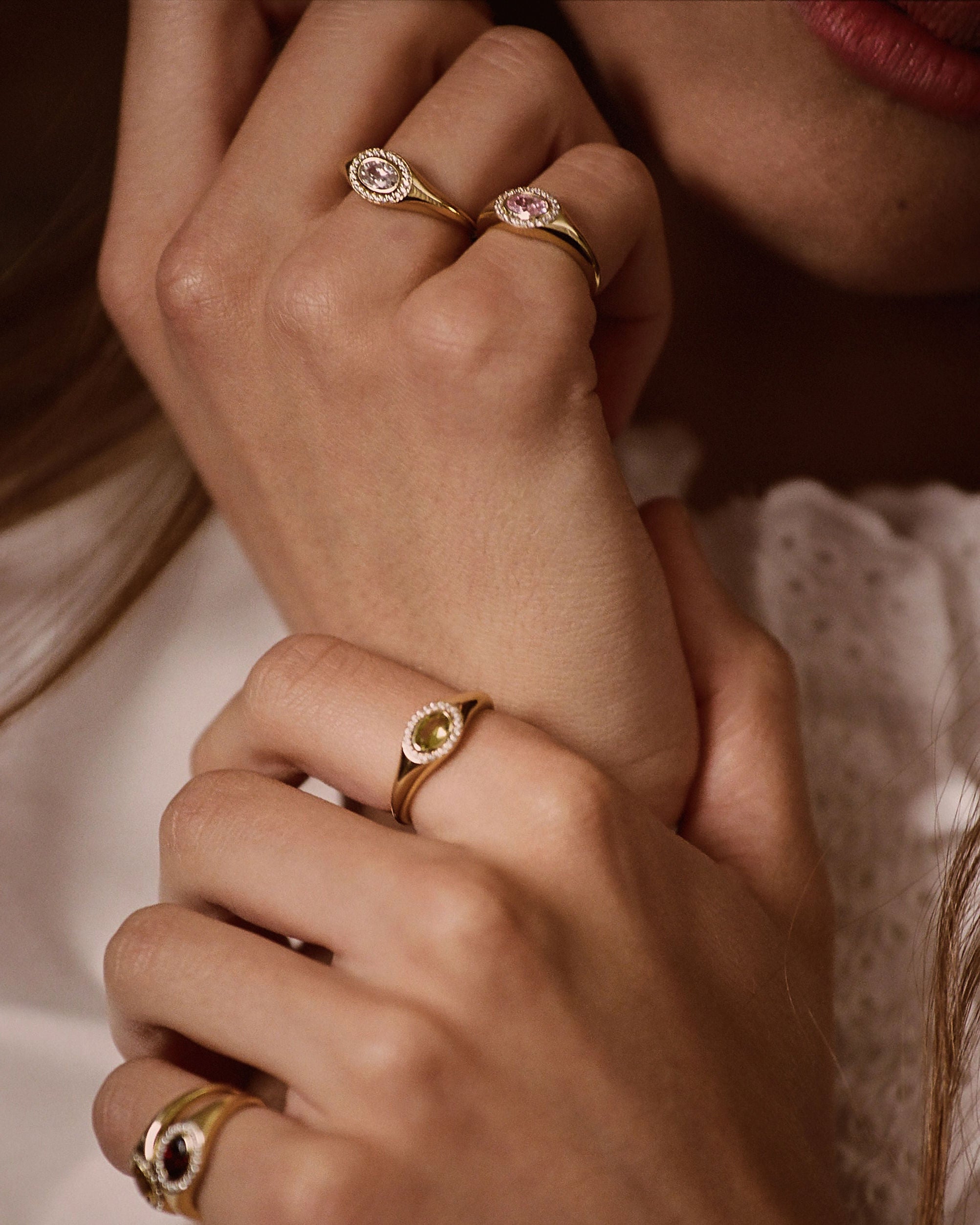 Close-up of a person wearing multiple gold rings with oval gemstones, including one with a pink stone and another with a dark stone, against a soft white lace garment.
