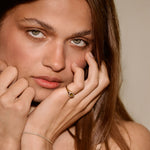 A close-up portrait of a woman with long brown hair wearing a delicate gold chain bracelet on one wrist and a small gold ring on one finger, posing with her hands near her face against a soft beige background.