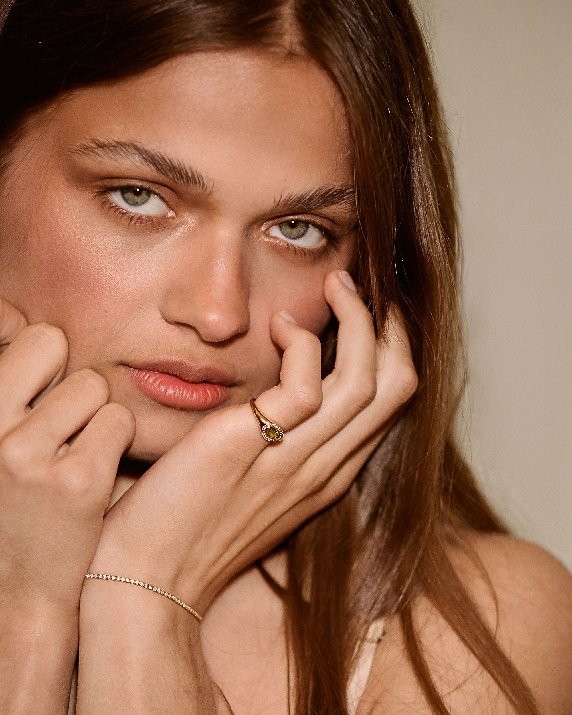 A close-up portrait of a woman with long brown hair wearing a delicate gold chain bracelet on one wrist and a small gold ring on one finger, posing with her hands near her face against a soft beige background.