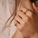 Close-up of a person's hands wearing gold rings, including one ring with a crescent moon and star design and another with a dark oval stone, set against a soft beige background.