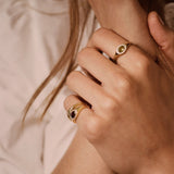 Close-up of a person's hands wearing gold rings, including one ring with a crescent moon and star design and another with a dark oval stone, set against a soft beige background.