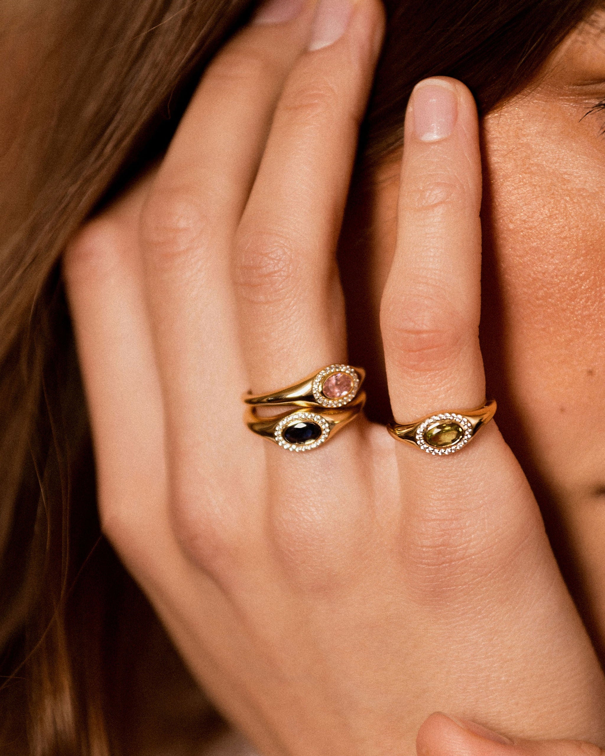 Close-up of a person's hand wearing two gold rings with oval gemstone settings, one with a pink stone and one with a dark stone, accented by small clear stones.