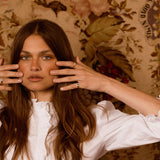 A woman with long brown hair poses in front of a vintage floral wallpaper, wearing a white ruffled blouse and holding her hands near her face.